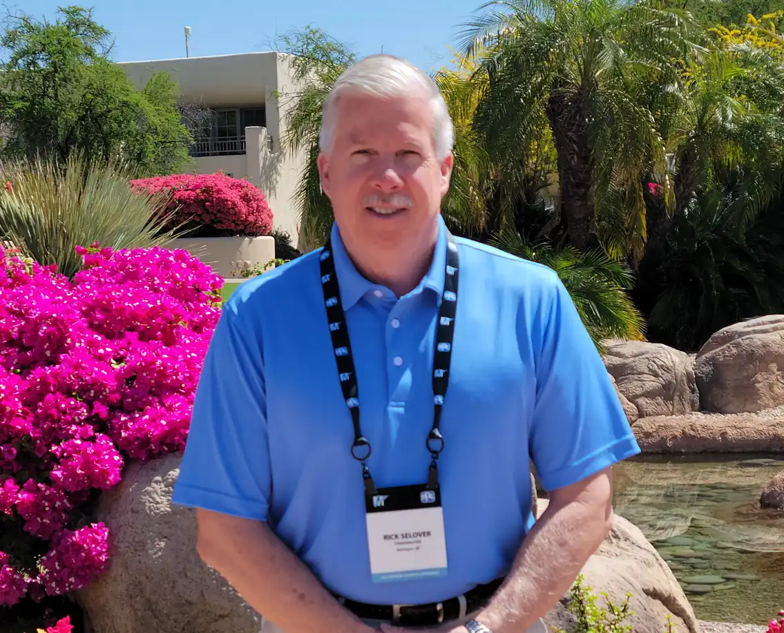 A middle-aged man with short, white hair is smiling at the camera. He's wearing a blue polo shirt and a conference badge around his neck with the name "RICK SELOVER". He's standing outdoors in a landscaped garden area with bright pink bougainvillea bushes, palm trees, and rocks visible in the background.