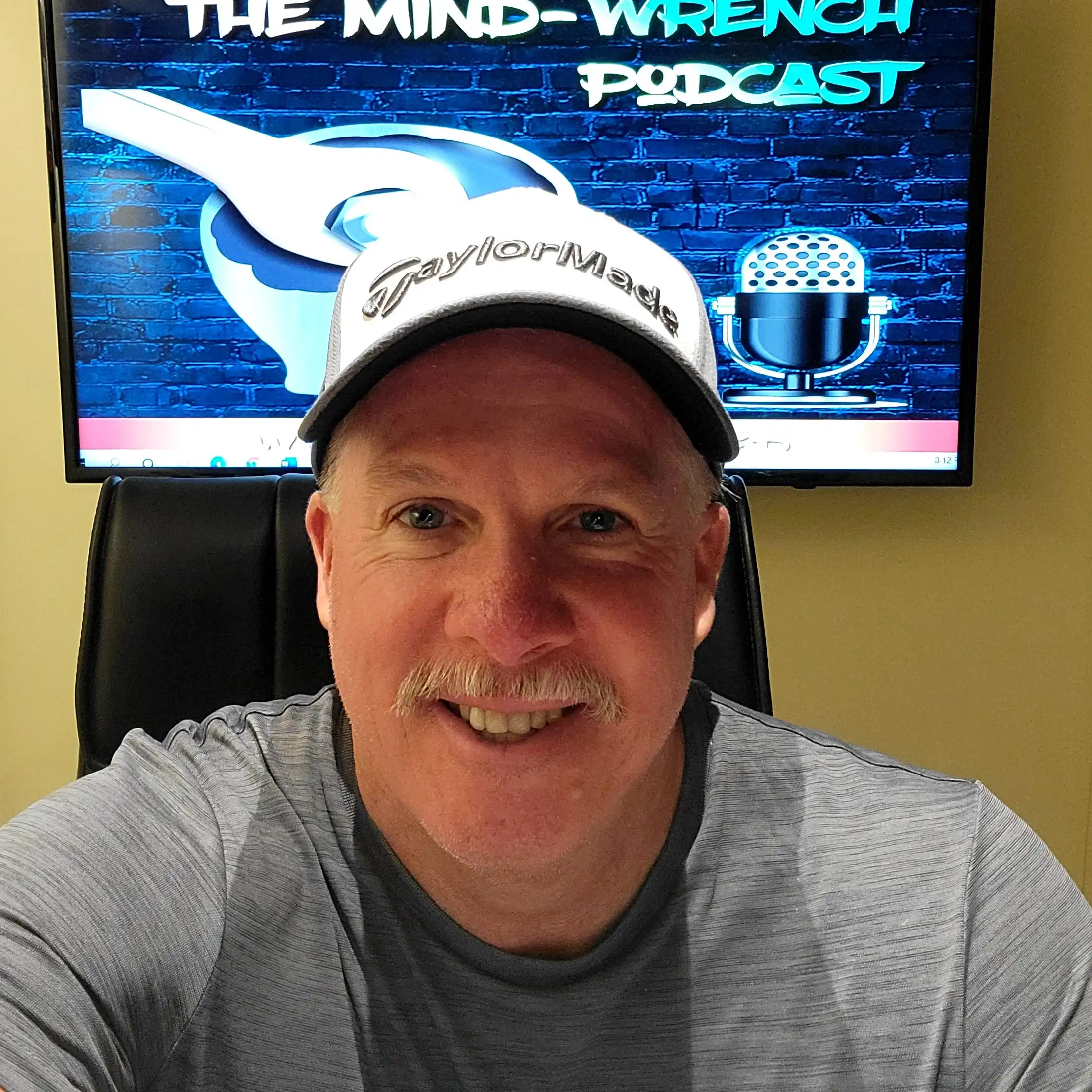 Close-up selfie of Rick Selover wearing a grey shirt and TaylorMade cap in front of a screen displaying The Mind-Wrench Podcast logo.