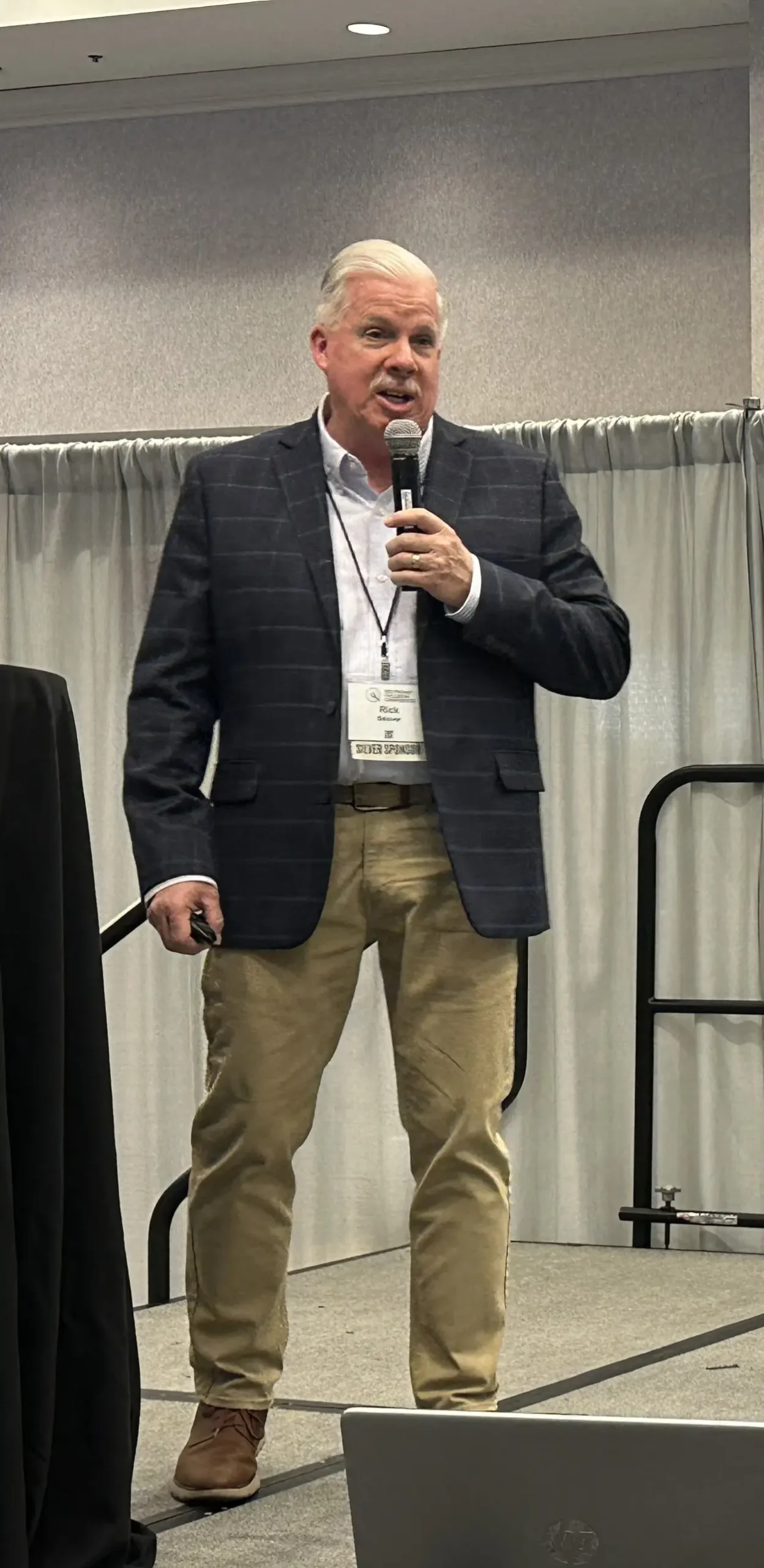 Rick Selover standing on stage holding a microphone during a conference presentation in a navy plaid blazer and khaki pants.